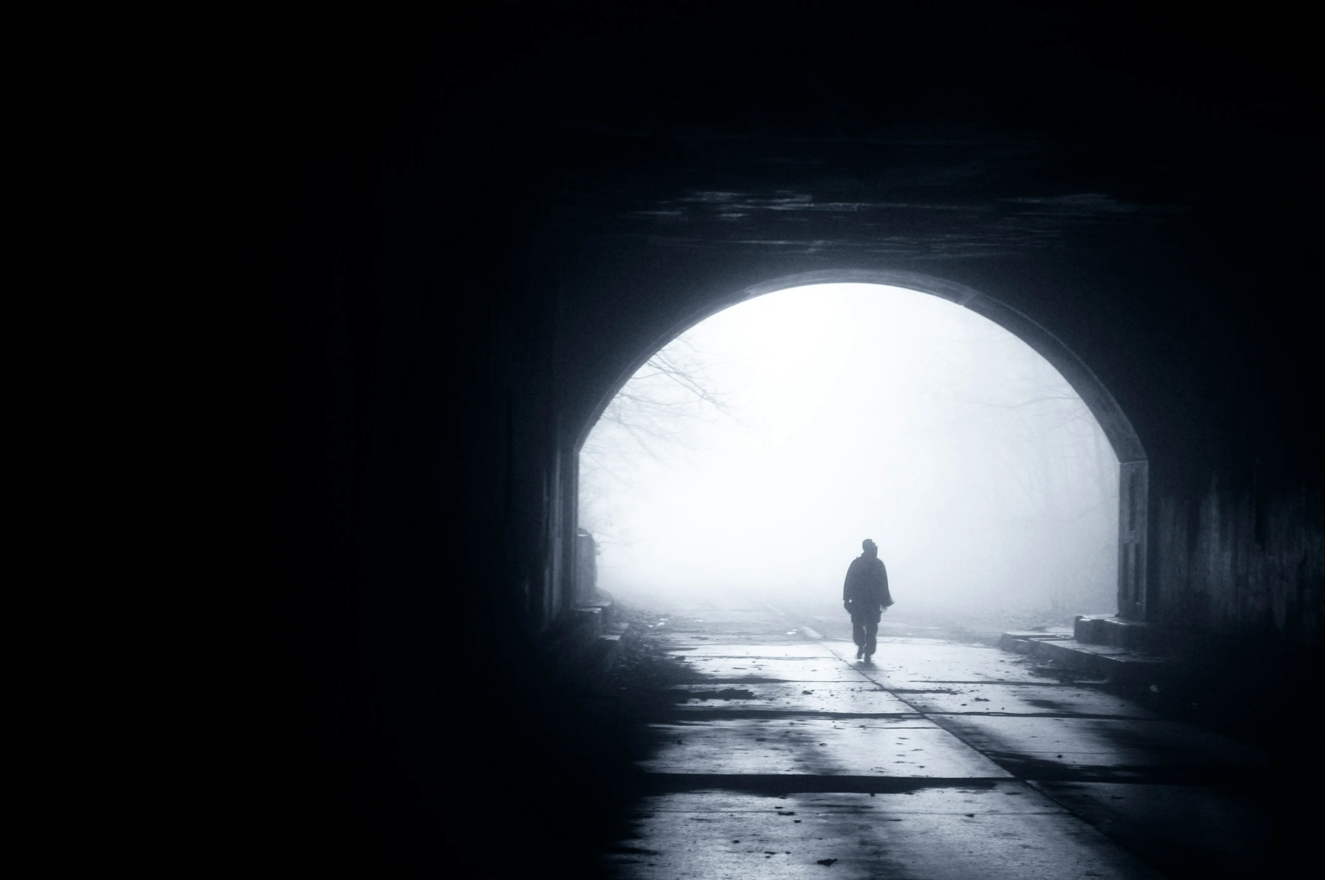 Silhouette of a lone person walking out of a dark tunnel towards the bright, misty opening
